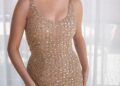 A woman in a sparkling gold dress poses elegantly by a sheer curtain.
