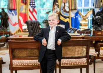 A young boy in a suit smiles, leaning against two chairs in the White House.