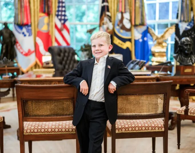 A young boy in a suit smiles, leaning against two chairs in the White House.