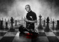 A man stands on a large chessboard with oversized pieces. A fallen king piece lies near him with a red stain beneath it.