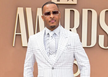 T.I. in a plaid suit and sunglasses stands on a red carpet at an awards event.