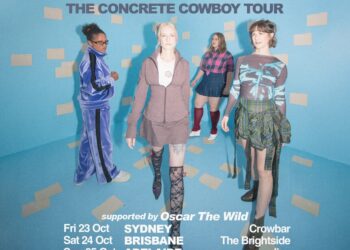 Four women stand in the corner of a blue room with grey rectangle patterns, white text detail tour