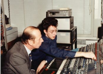 Two men collaborating at a sound mixing console, adjusting sliders and settings in a recording studio environment.