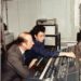 Two men collaborating at a sound mixing console, adjusting sliders and settings in a recording studio environment.