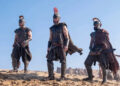 Jimmy Gonzales, Matt Damon and Himesh Patel in a scene from "The Odyssey." Three men in Greek soldier dress an armor stand on a dirt battlefield.