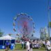People walk among vendor booths and carnival rides on a sunny day at the San Joaquin County Fairgrounds