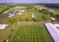 An aerial view of Trinity Park in Ipswich. The showground is set up for the Suffolk Show, but people are yet to be let in. We can see several arenas with patterned, mown grass as well as temporary white tents or varying sizes, roadways, and some trees. The rest of the countryside is visible on the horizon, as are the cranes at the Port of Felixstowe.