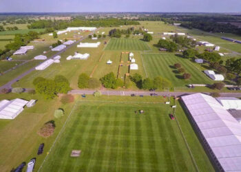 An aerial view of Trinity Park in Ipswich. The showground is set up for the Suffolk Show, but people are yet to be let in. We can see several arenas with patterned, mown grass as well as temporary white tents or varying sizes, roadways, and some trees. The rest of the countryside is visible on the horizon, as are the cranes at the Port of Felixstowe.