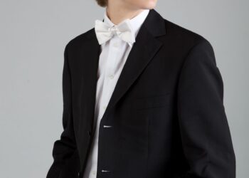 Teen boy wearing a black suit jacket, white shirt, and white bow tie, standing with hands in pockets, looking to the side against a plain background.