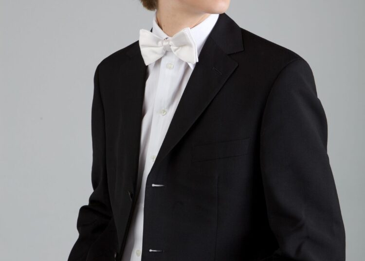 Teen boy wearing a black suit jacket, white shirt, and white bow tie, standing with hands in pockets, looking to the side against a plain background.
