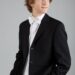 Teen boy wearing a black suit jacket, white shirt, and white bow tie, standing with hands in pockets, looking to the side against a plain background.