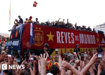 An open-top bus carries the Spain national football team through the streets of Madrid. Crowds cheer in celebration from the street.
