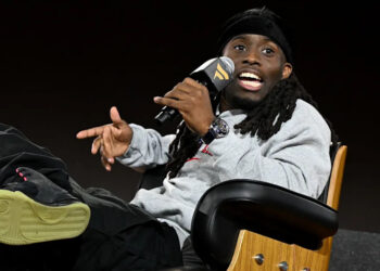 Kai Cenat with long hair and a cap, wearing a gray sweatshirt and black pants, sits in a chair speaking into a microphone.