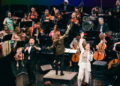 A man in a black tuxedo and a woman in a white suit sing while an orchestra plays behind them.