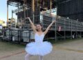 A young woman in a white tutu, white tights, and white socks over pointe shoes poses in back of a large outdoor stage. She pops her right foot up onto pointe and throws her arms up, palms flexed, and wears a large, enthusiastic smile.