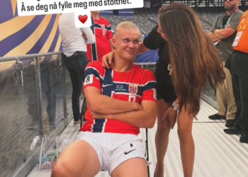 A young man in a Norway soccer jersey sitting while a woman interacts with him in a stadium setting.