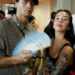 A young man holding a fan and a woman with a tattoo, both posing in a crowded indoor setting.