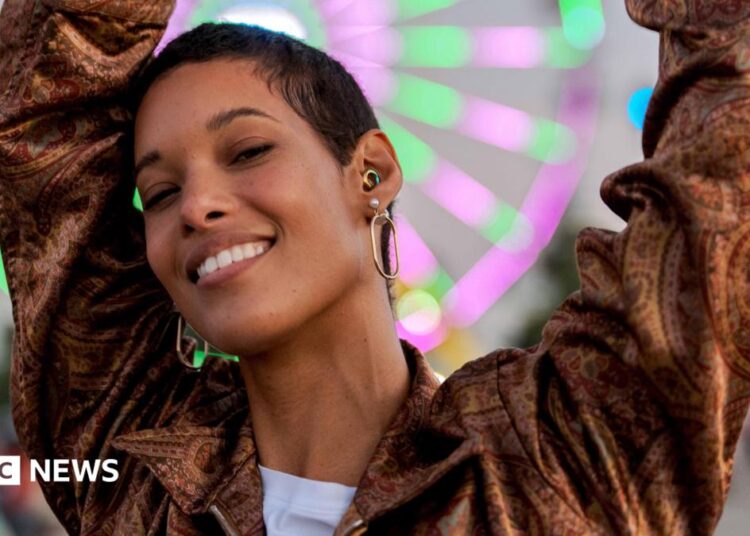 A young woman dances at dusk in front of a neon-lit Ferris wheel at a music festival. As well as large hoop earrings, she wears a rainbow-coloured ear-plug as protection for her hearing.