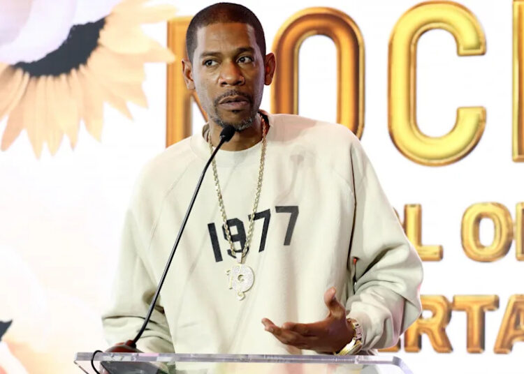 Young Guru in a beige sweatshirt with "1977" printed on it speaks at a podium. The background features floral designs and large text.