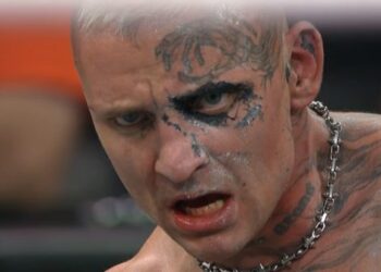 A close-up of a wrestler with a fierce expression, displaying striking facial tattoos and body art. The wrestler has a chain around his neck and appears to be in a high-stakes moment after a match.