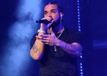 Drake actuando en el escenario, sosteniendo un micrófono, vistiendo una camisa negra y joyas, con un fondo iluminado en azul.