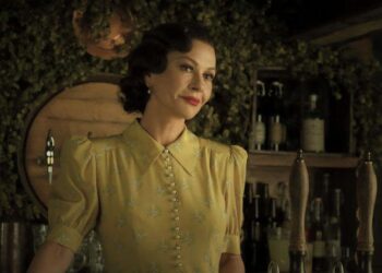 Catherine Zeta Jones con un vestido amarillo. Tiene el pelo corto y oscuro y está parada detrás de una barra de estilo antiguo.