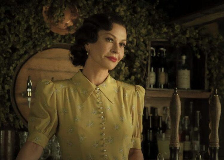 Catherine Zeta Jones con un vestido amarillo. Tiene el pelo corto y oscuro y está parada detrás de una barra de estilo antiguo.
