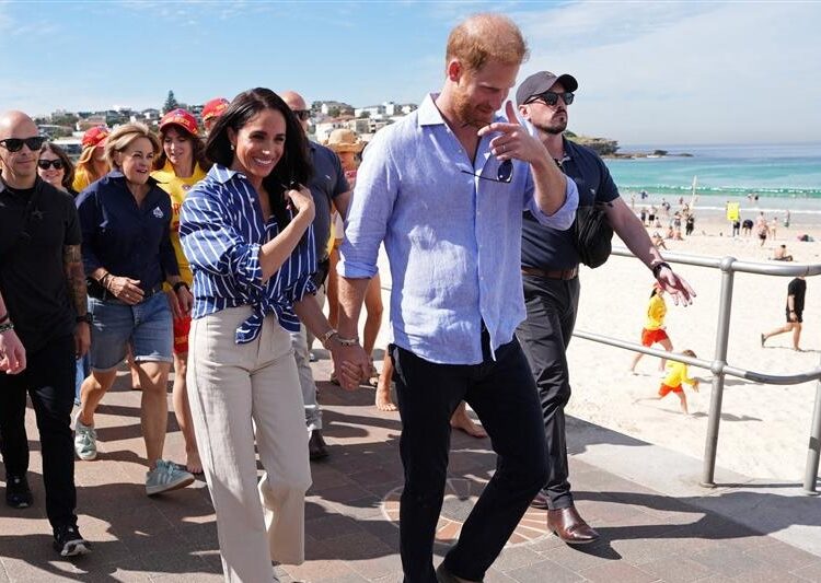 Harry y Meghan caminan por Bondi Beach con salvavidas de surf detrás de ellos.