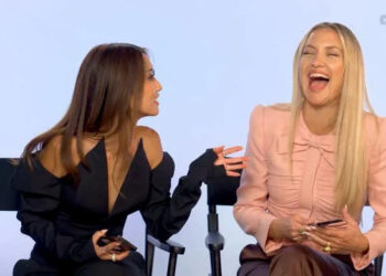 Dos mujeres sentadas y riendo; uno con un elegante conjunto negro con hombros descubiertos, el otro con una elegante blusa rosa con un lazo