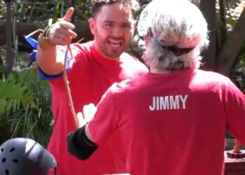 Dos individuos vestidos con camisetas rojas, uno con un visible "PALANQUETA" impreso en la parte posterior, están juntos en un ambiente al aire libre, y uno de ellos hace un gesto con el dedo.