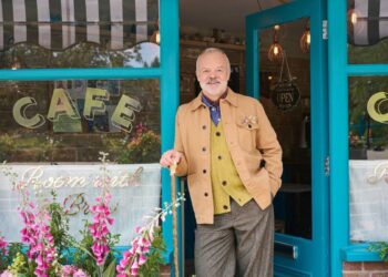 Un señor mayor está de pie frente a la entrada de un color azul vibrante de una cafetería, vestido con una chaqueta color canela y pantalones grises. El exterior del café está adornado con flores de colores y tiene una puerta de cristal con la palabra "CAFETERÍA" exhibido de manera destacada en letras iluminadas.