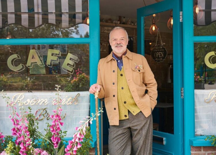 Un señor mayor está de pie frente a la entrada de un color azul vibrante de una cafetería, vestido con una chaqueta color canela y pantalones grises. El exterior del café está adornado con flores de colores y tiene una puerta de cristal con la palabra "CAFETERÍA" exhibido de manera destacada en letras iluminadas.