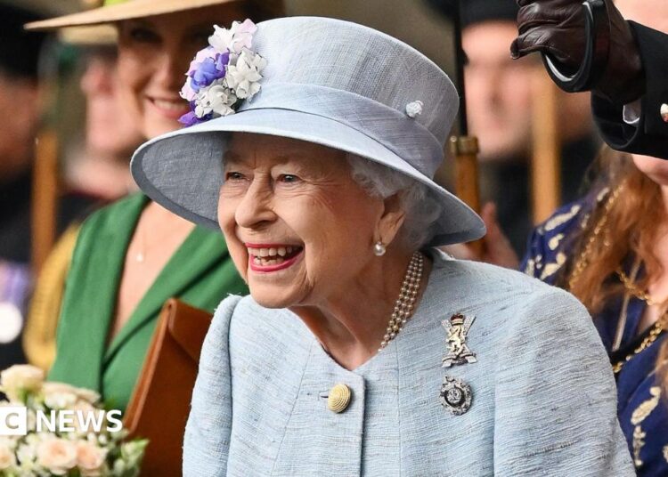 King Charles III with his late mother, Queen Elizabeth II - both are smiling off camera, and standing on the Buckingham Palace balcony which can just be seen. The late Queen wears an emerald green hat and jacket, with pearls and a diamond brooch in the shape of a bow, while the King has a navy pin-striped suit and a pale blue patterned tie