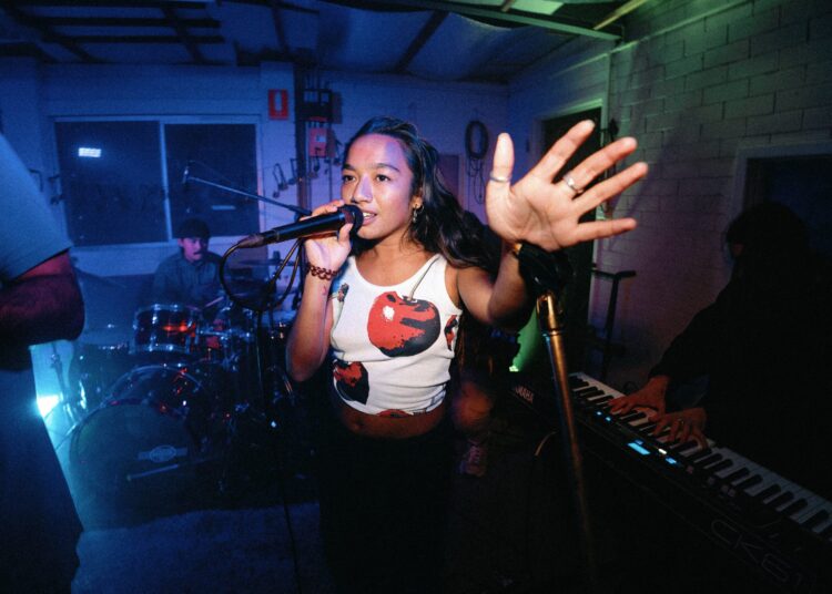A woman sings into a microphone with her hand raised, performing with a band. A drummer and keyboard player are visible in the background under colorful stage lights.