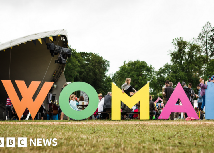 A field with a large music stage in the background and lots of people in front of it. In the foreground of the picture there are huge multi-coloured letters spelling out 'WOMAD' in capital letters