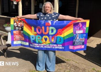 A woman stands outdoors holding a colourful “Loud & Proud” Pride festival banner in front of a wooden building with hanging baskets and string lights.