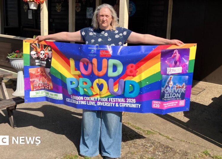 A woman stands outdoors holding a colourful “Loud & Proud” Pride festival banner in front of a wooden building with hanging baskets and string lights.
