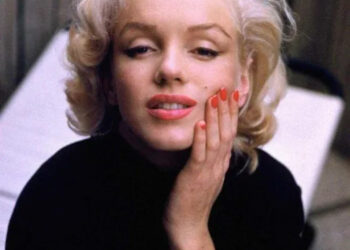 A woman with short, wavy blonde hair and red lipstick rests her hand on her cheek, wearing a black top and red nail polish, looking slightly up with a gentle expression. A blurred background is visible.