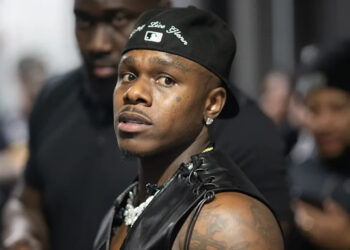 DaBaby con una gorra y un chaleco negros, mirando hacia un lado, con una multitud borrosa al fondo.
