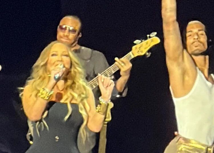 Mariah Carey, cantando frente a un micrófono, mientras Alex Jackson está a su derecha bailando, con un brazo levantado en el aire.