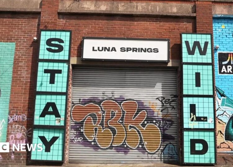 A grey metal shutter with orange graffiti is pulled over the entrance to Luna Springs. Murals are painted on the red brick wall around its entrance. Signs either side of the entrance say 'Stay Wild' in blue, with a white 'Luna Springs' sign above.