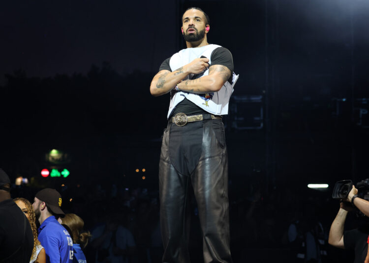 Drake performing on stage, wearing a black shirt and leather pants, with a crowd and security in the background.