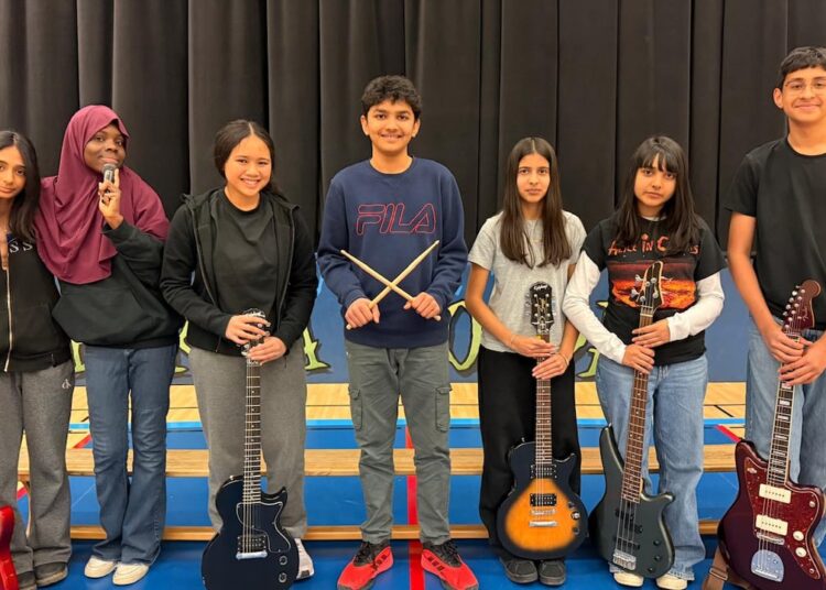 Los niños en una clase de música posan con instrumentos frente a un escenario con una cortina negra.