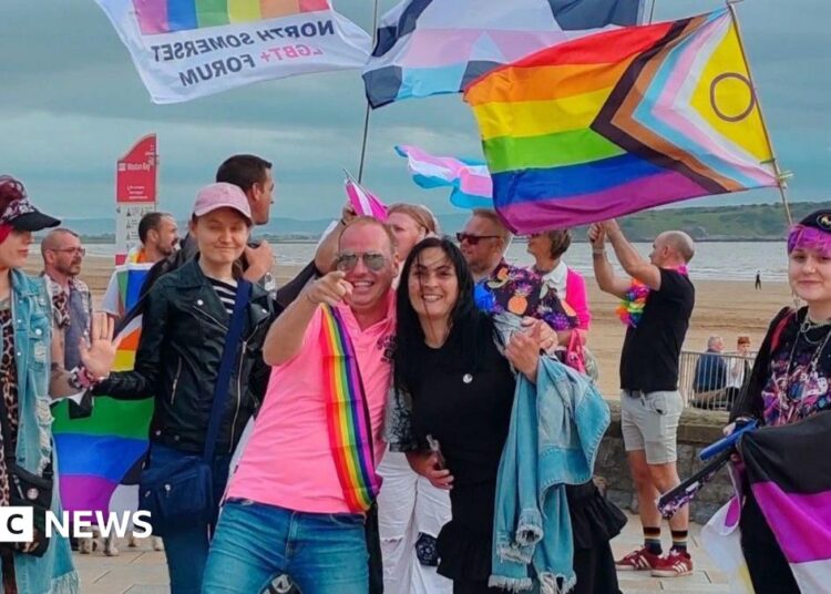 A group of people dressed in different colours and waving LGTBQ flags are standing by the beach in Weston-super-Mare.  Several people are smiling towards the camera.