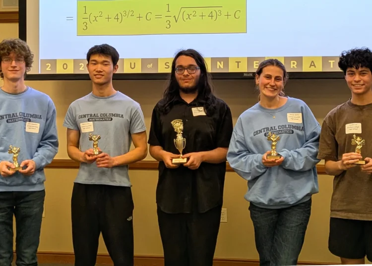 Cinco estudiantes posan en el interior sosteniendo trofeos, con una ecuación matemática proyectada encima de ellos.