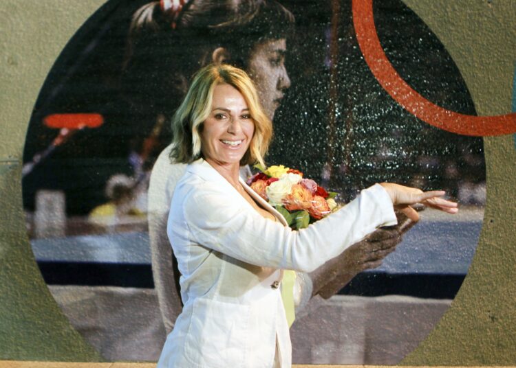 Nadia Comaneci smiles while posing in front of a large photo of her younger self.