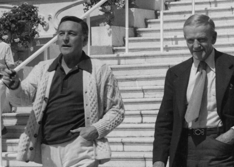Una imagen en blanco y negro de Gene Kelly y Fred Astaire bajando unas escaleras en Cannes. Kelly sostiene un par de gafas de sol y lleva una camiseta polo oscura con un cárdigan de punto grueso. Astaire lleva traje oscuro y corbata. Ambos hombres tienen una mano en el bolsillo.