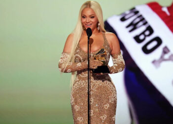 Beyoncé, con un vestido dorado brillante, sostiene un premio Grammy mientras habla frente a un micrófono en el escenario.