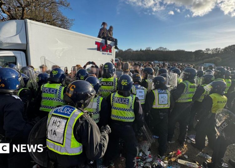 White truck with two people sat on the roof - dozens of police officers in riot gear are standing in a group with a large crown seen beyond in a field.