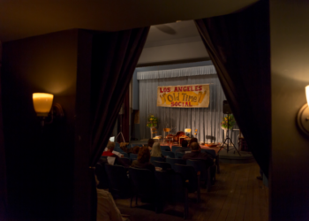 Una vista de un pequeño escenario con un cartel que dice "Social de antaño en Los Ángeles.' Algunas personas se sientan en sillas entre el público.